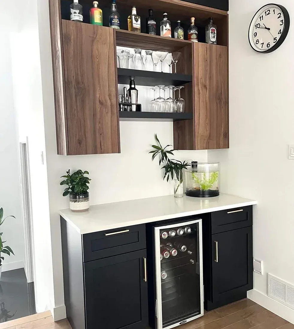 Bar à domicile avec armoires noires, étagères en bois et comptoir blanc ; comprend un réfrigérateur à vin, des verres et des bouteilles d'alcool.