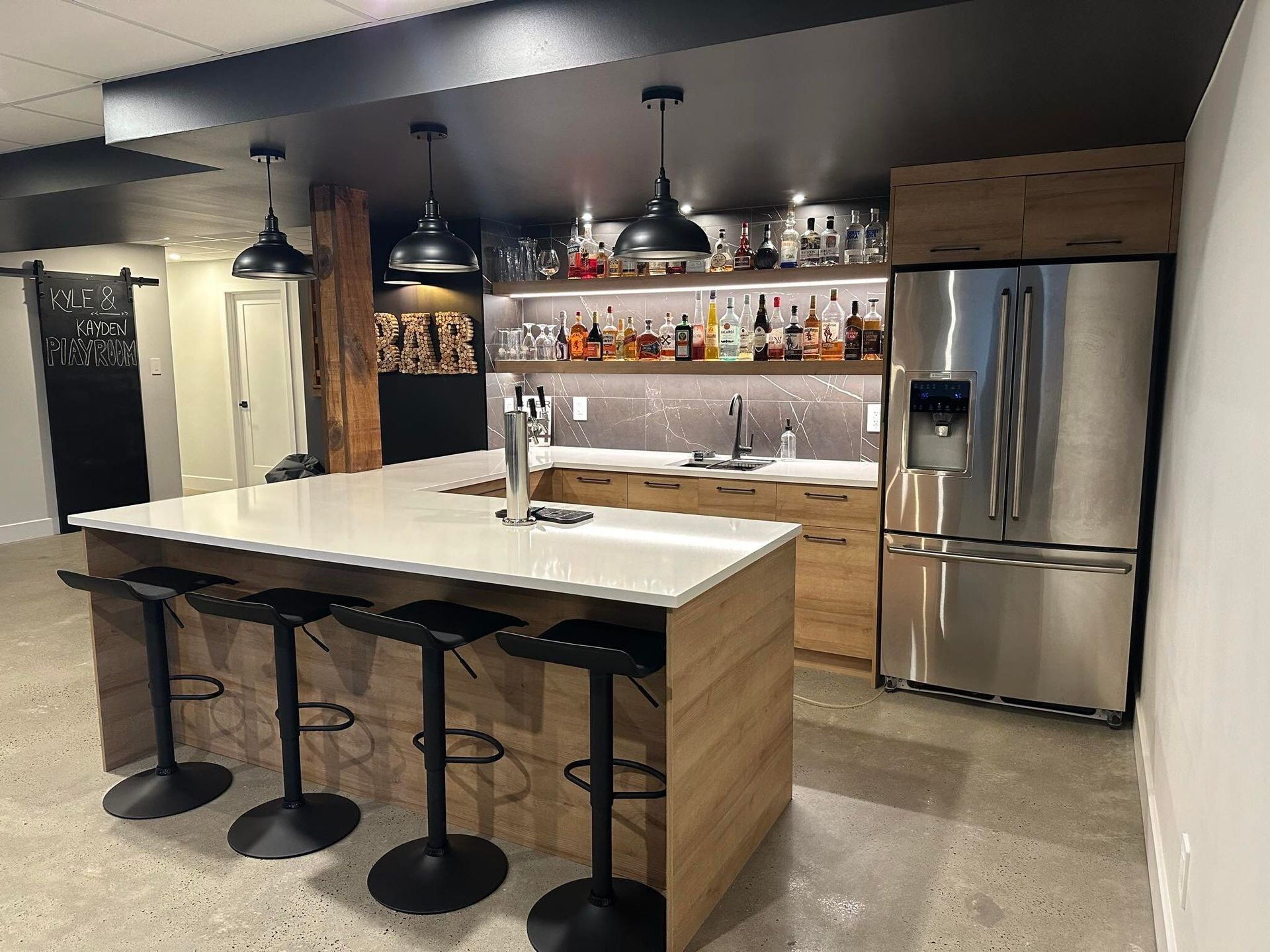 Bar au sous-sol avec îlot central, présentoir à boissons alcoolisées et réfrigérateur en acier inoxydable.