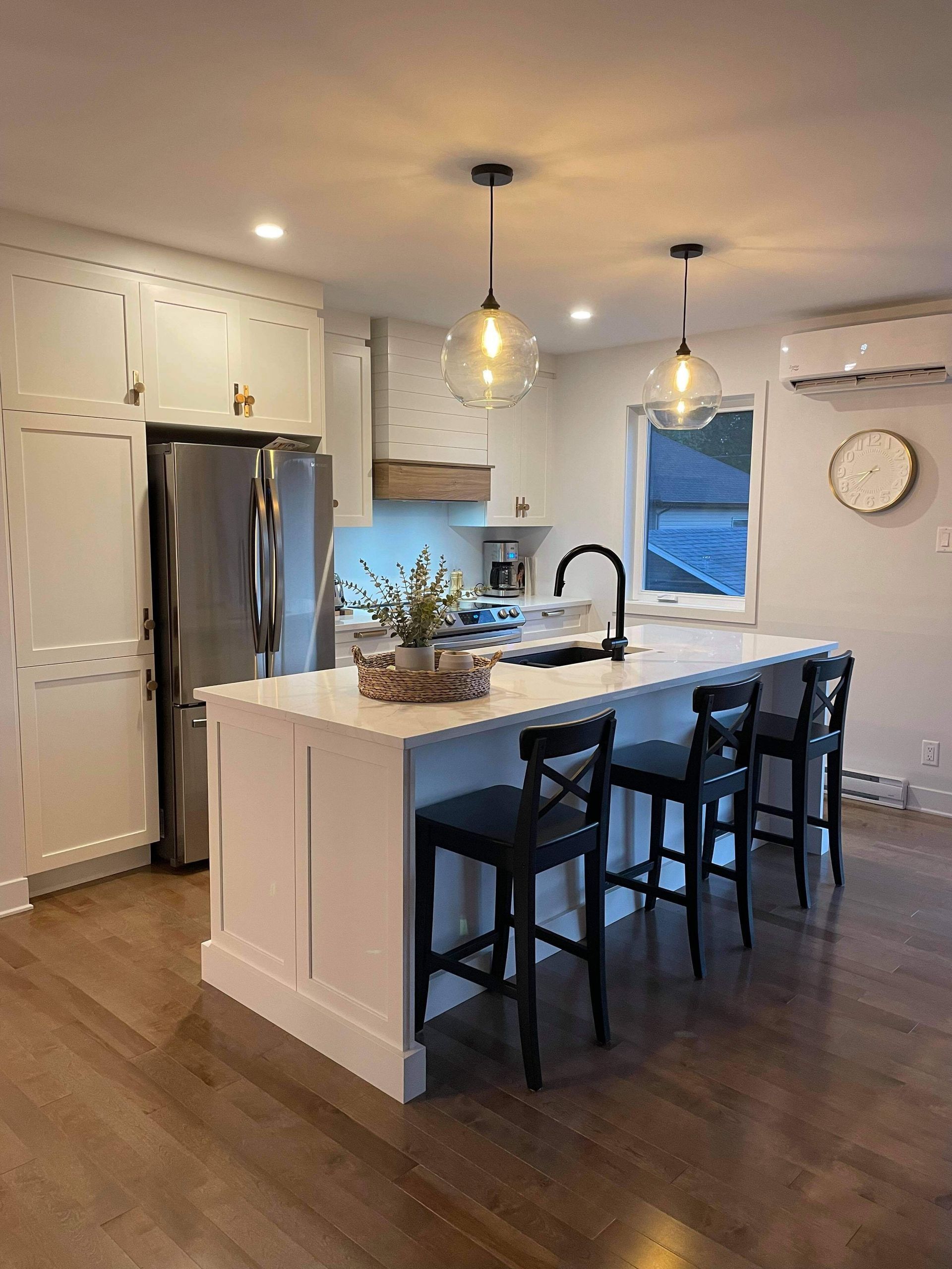 Cuisine moderne avec armoires blanches, îlot, tabourets noirs, réfrigérateur en acier inoxydable et deux luminaires suspendus.