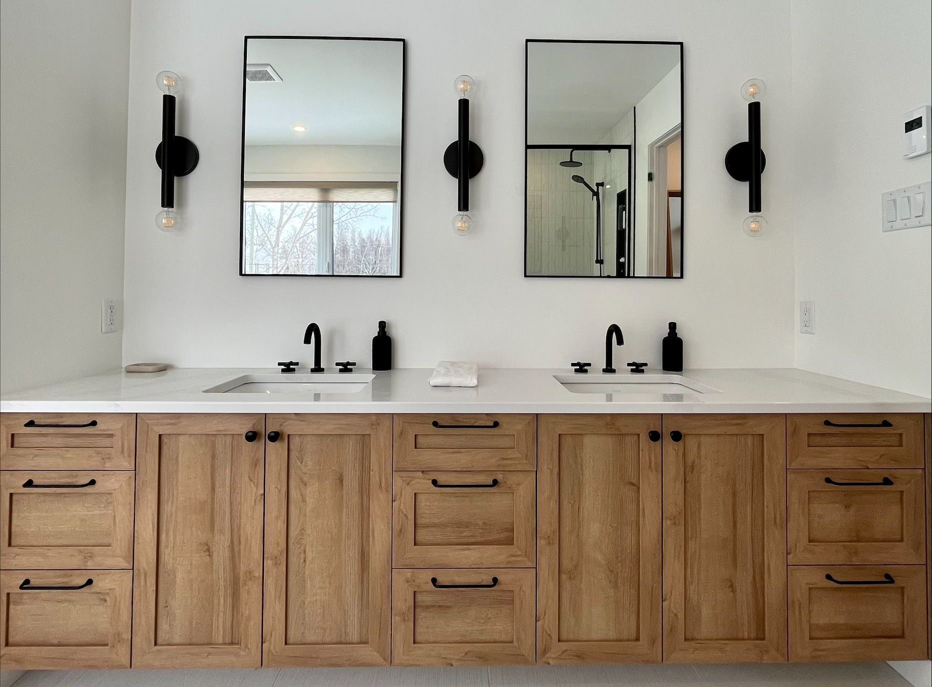 Salle de bain avec double vasque, deux miroirs noirs et appliques. Meubles en chêne avec plan de travail blanc.