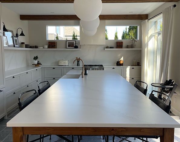 Cuisine moderne avec grand îlot blanc et table en bois. Armoires blanches et étagères ouvertes, baignées de lumière naturelle.