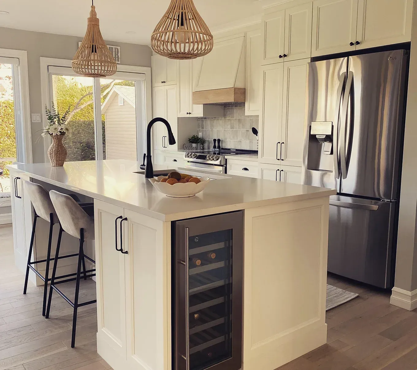 Cuisine blanche avec îlot central, cave à vin et réfrigérateur en acier inoxydable. Deux suspensions tissées sont suspendues au plafond.