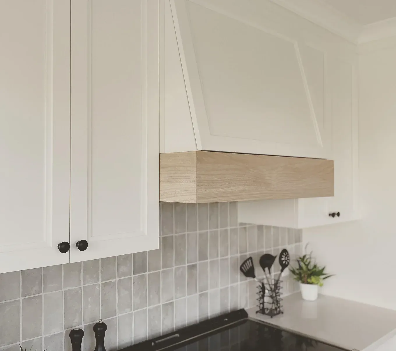 Armoires de cuisine blanches avec hotte garnie de bois, dosseret en carrelage gris et plaque de cuisson noire.