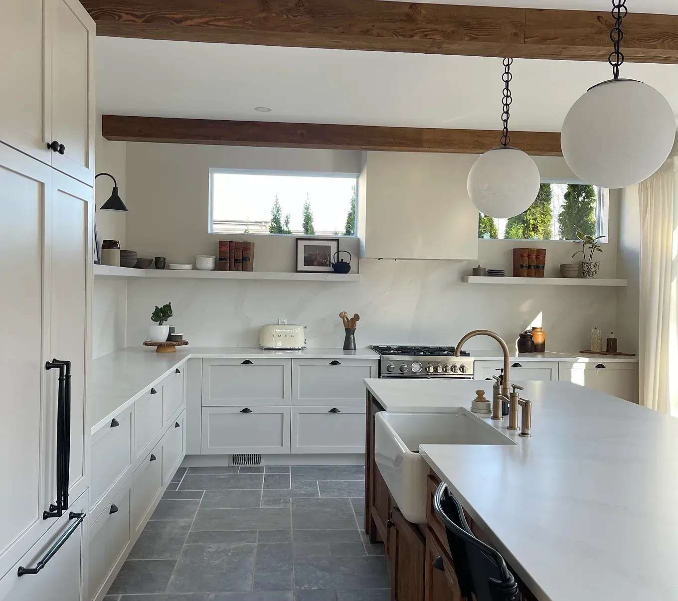 Cuisine moderne avec armoires blanches, îlot et sol en pierre; poutres en bois et lumières globe.