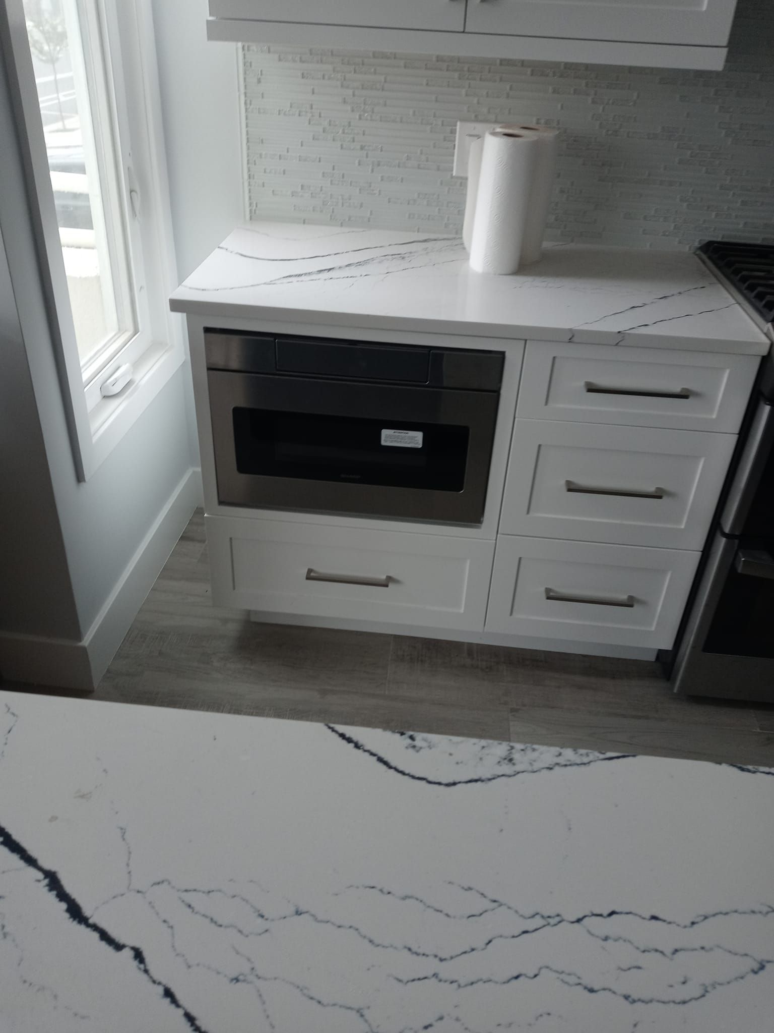 White kitchen cabinet with microwave and drawers, next to a stove, and window.