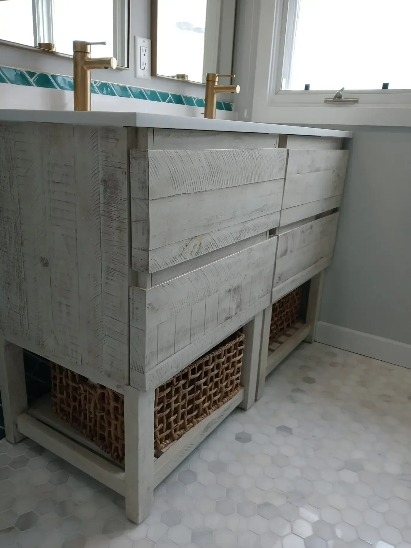 White-washed wooden double sink vanity with gold faucets, wicker baskets, and hexagon floor tiles.