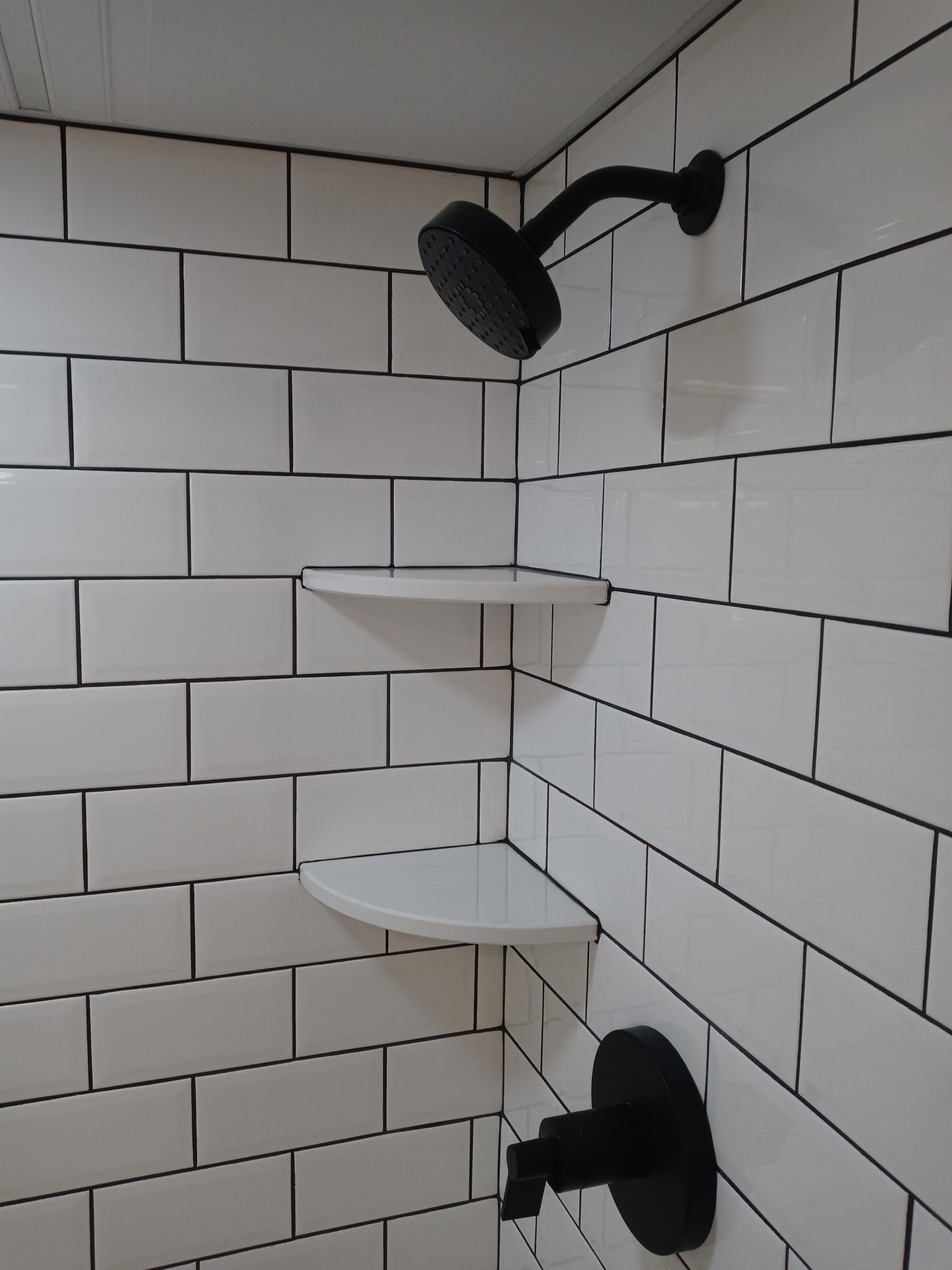 White tiled shower with black showerhead, shelves, and faucet.
