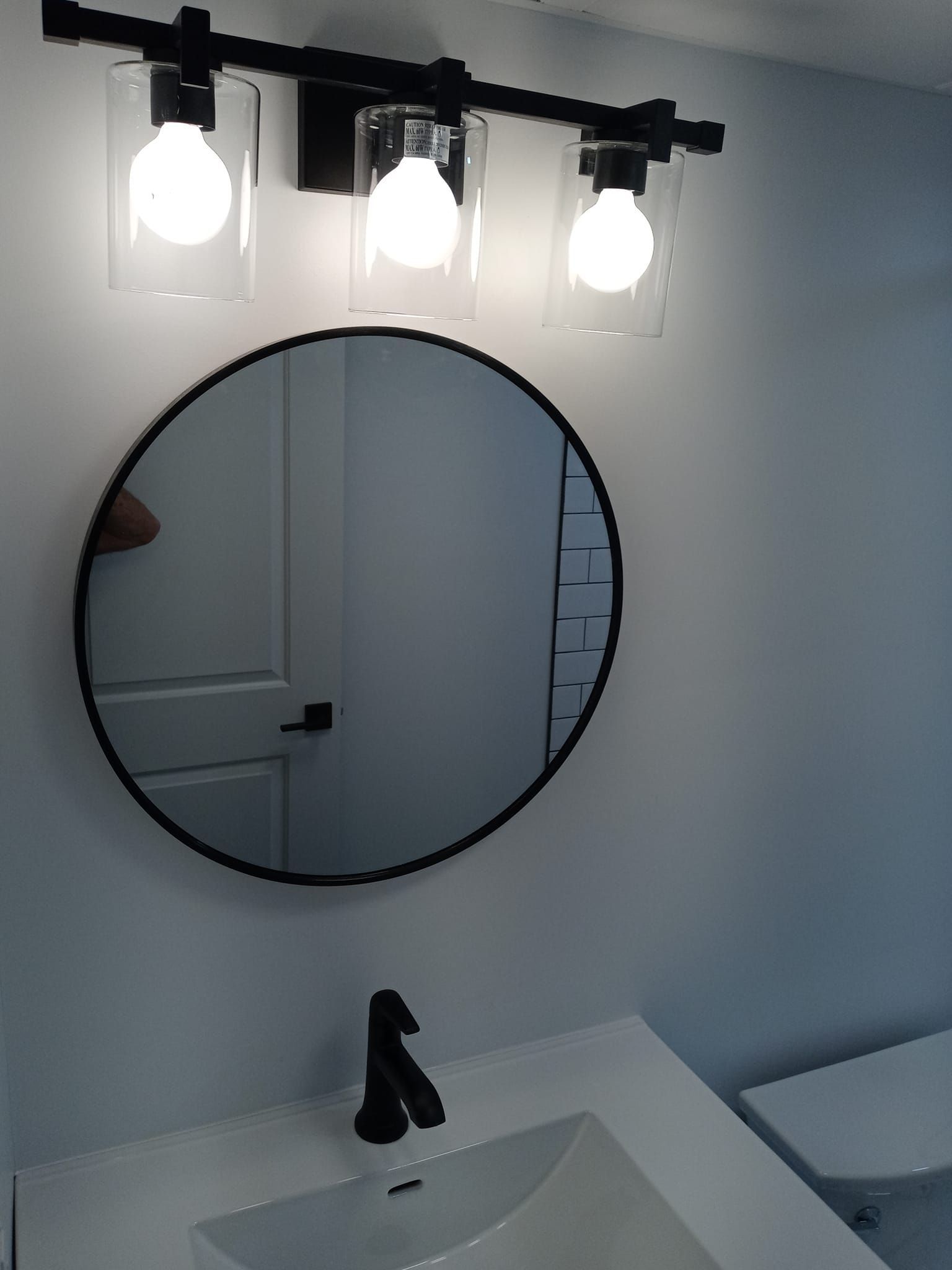 Bathroom vanity with round mirror, black faucet, and three-bulb light fixture over white sink.