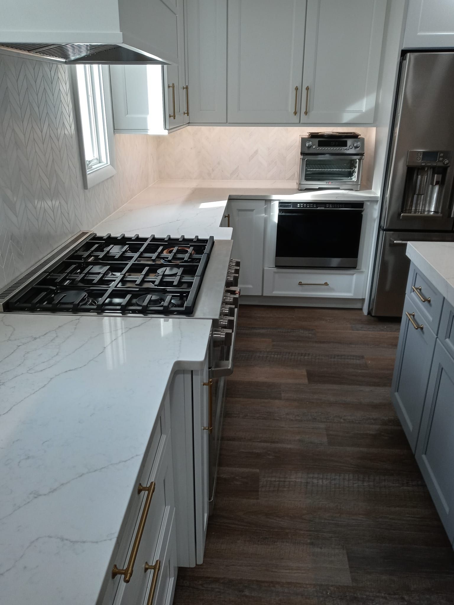 Modern white kitchen with gas range, oven, and stainless steel refrigerator. Wooden floor.