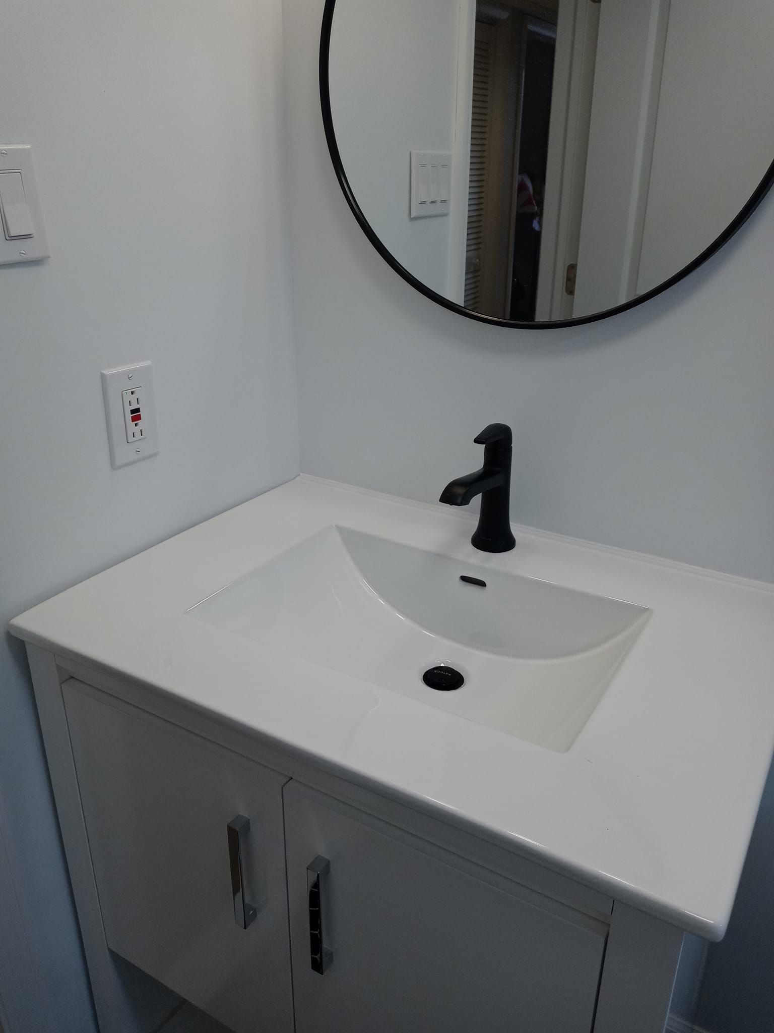 White bathroom sink with black faucet, mounted on white cabinet, with a round mirror above.