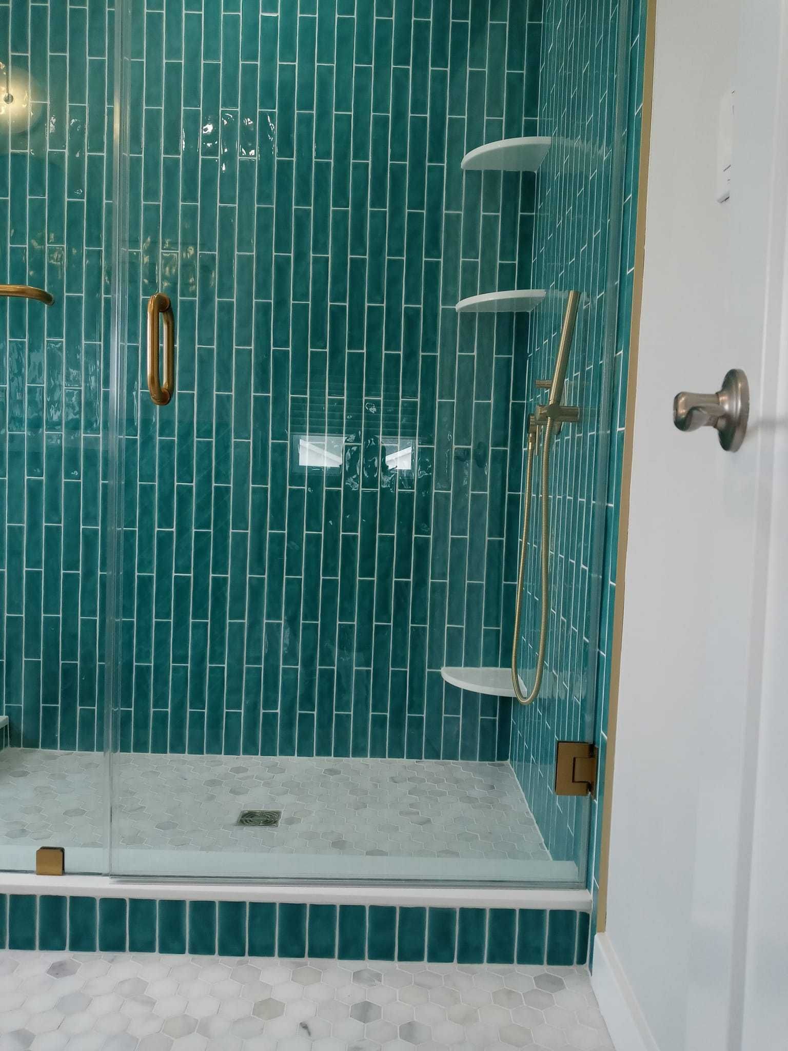 Teal tiled shower with glass door and gold fixtures. White shelves and grey stone floor.