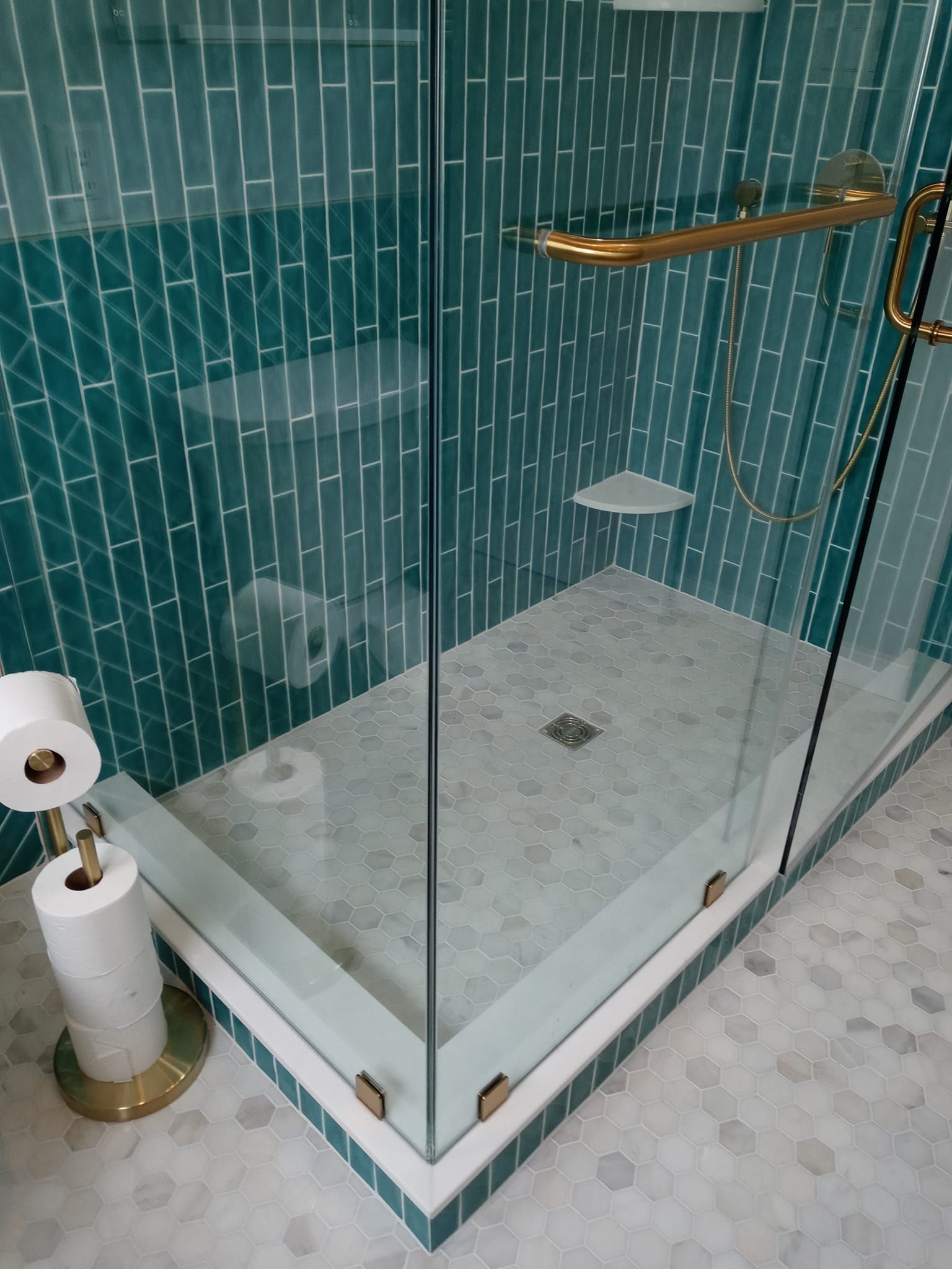 Teal tiled shower with glass doors, gold hardware, and a stone floor. A toilet paper holder is visible.