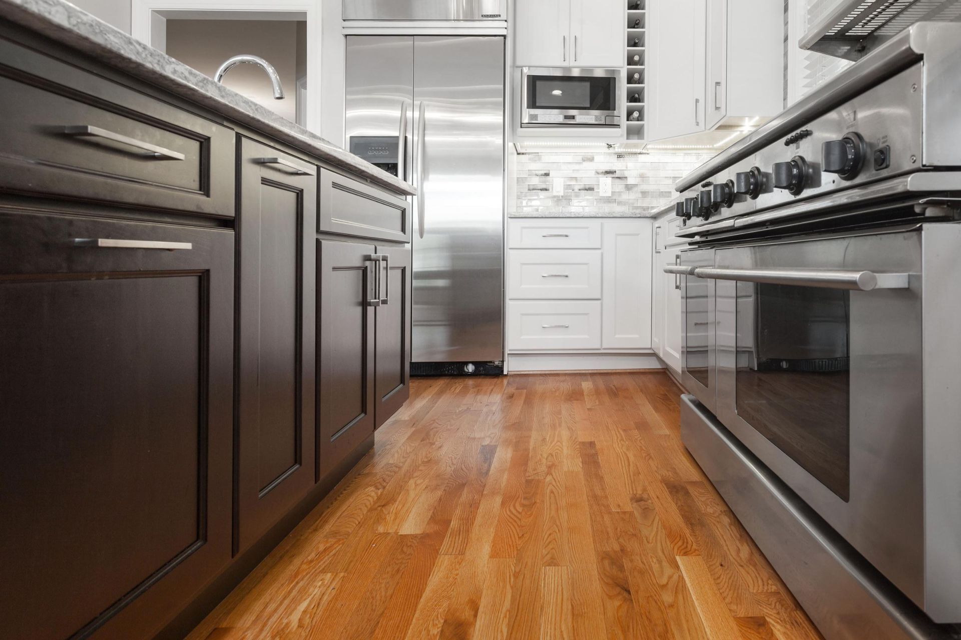 Wooden floor in a modern kitchen with dark brown and white cabinets, a stainless steel oven and refrigerator.