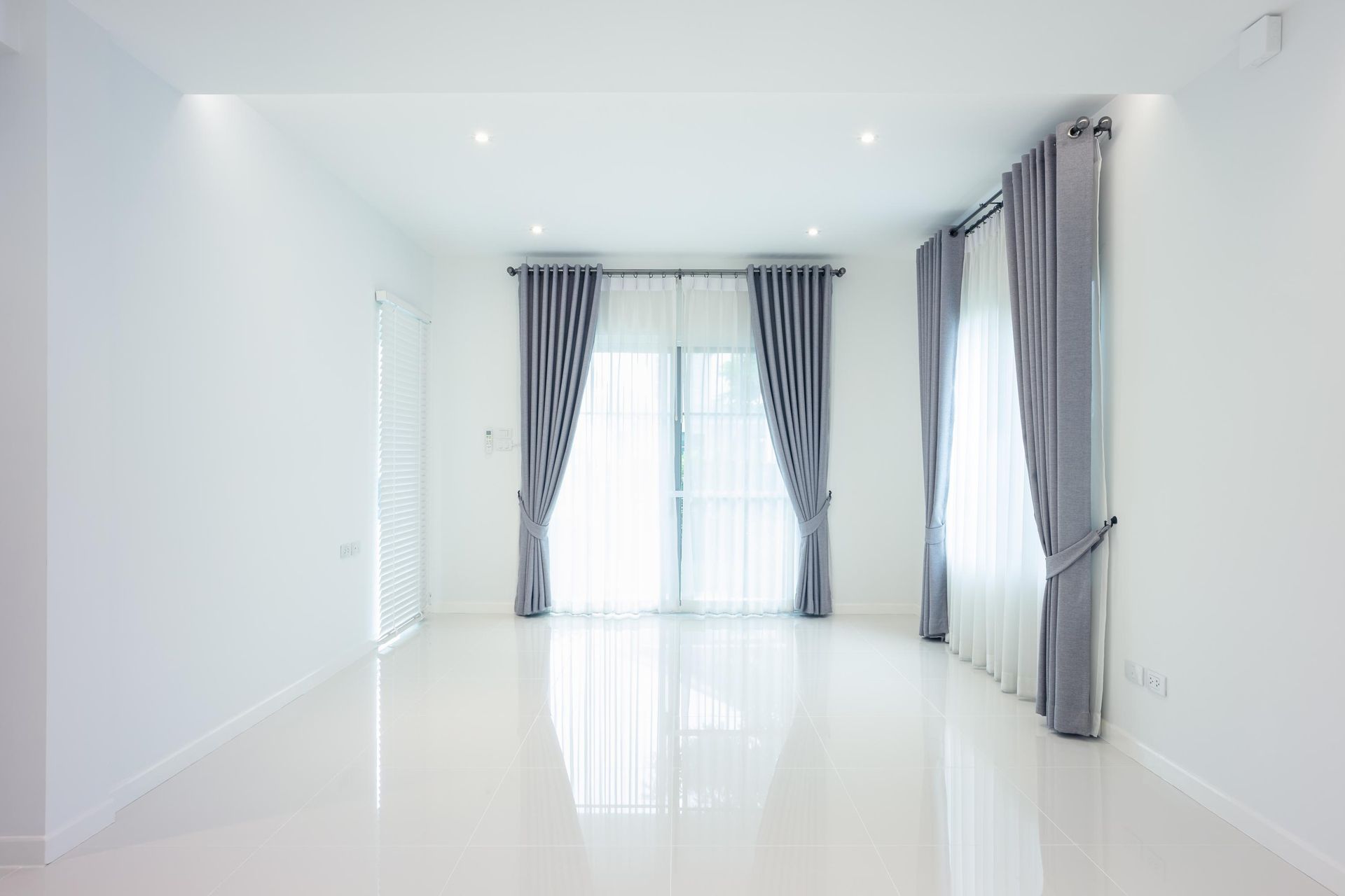 Empty white room with shiny floor, two windows with gray curtains, and recessed lighting.