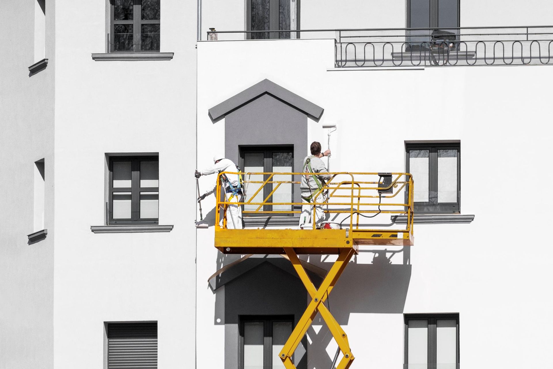 Two people painting a gray structure on a white building, using a yellow lift.