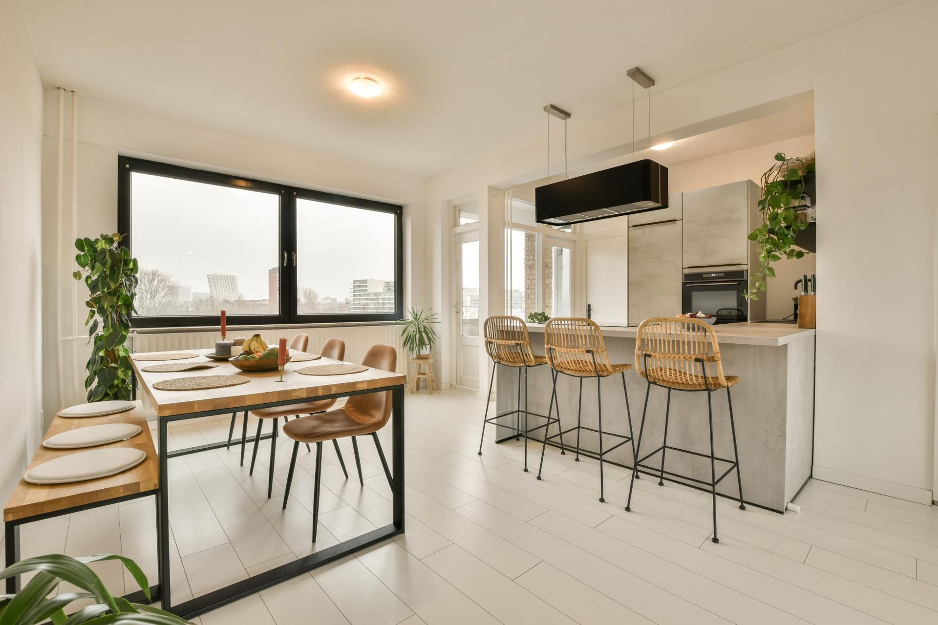 Modern dining room with table, chairs, bench, and kitchen island with bar stools. Bright, white space with plants.