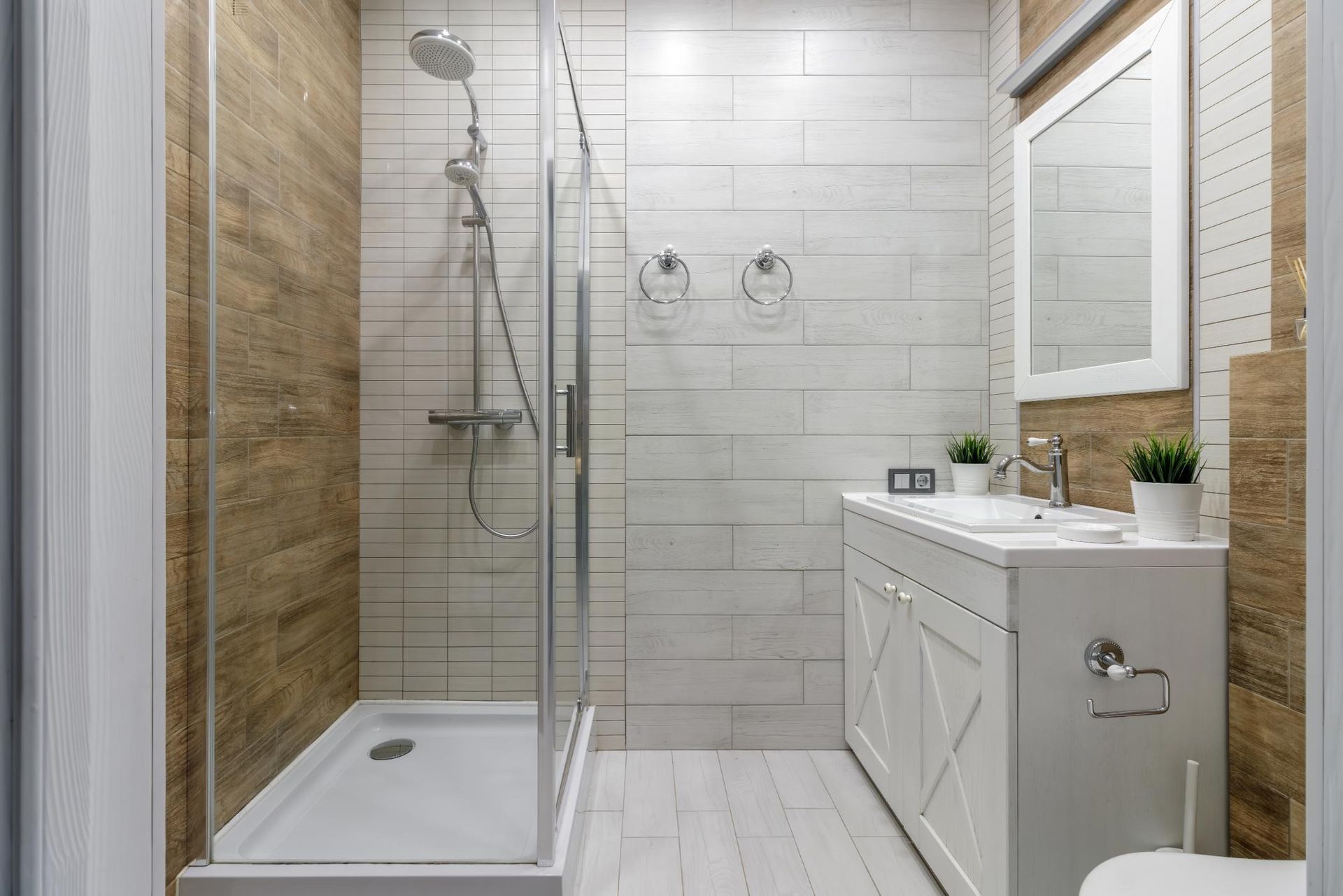 Small bathroom with white and wood-toned tiles, glass shower, white vanity, and toilet.