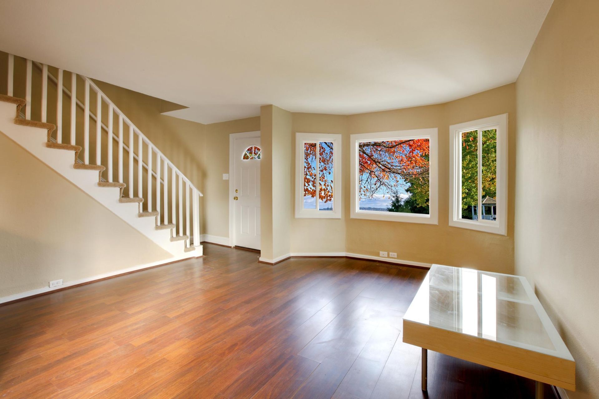 Empty, light-filled living room with hardwood floors, stairs, and three windows overlooking a tree.