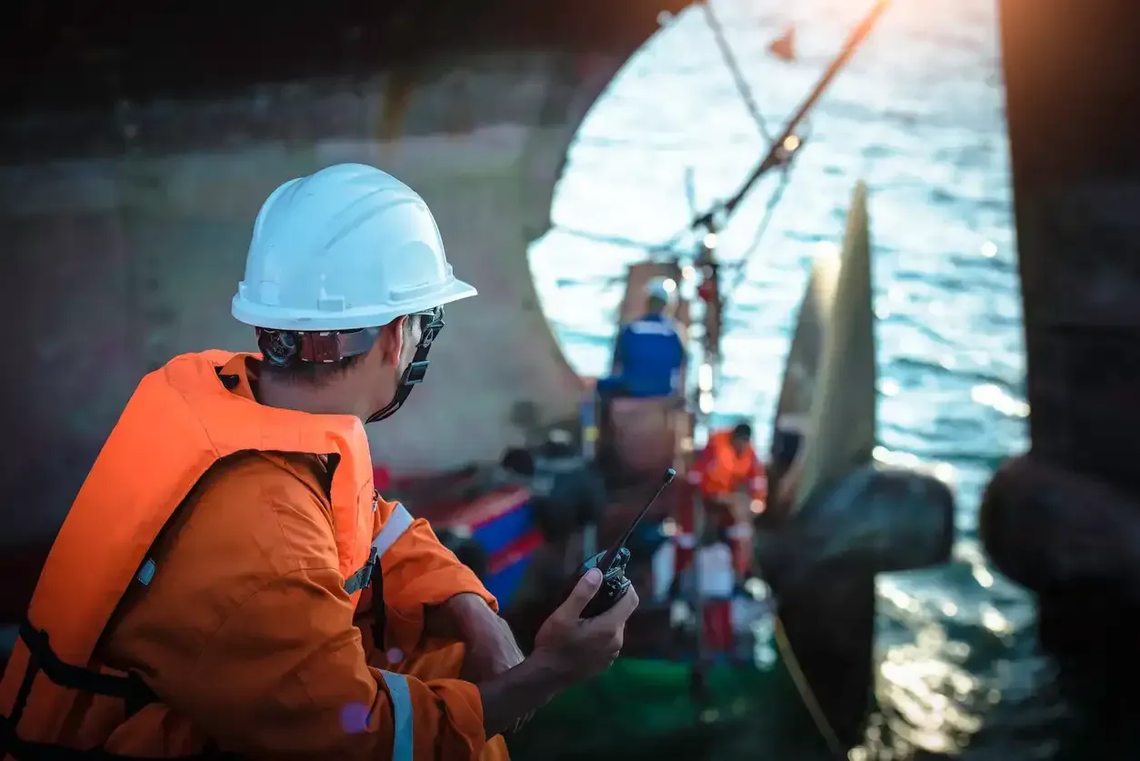 A man wearing a life jacket and a hard hat is using a cell phone on a boat.