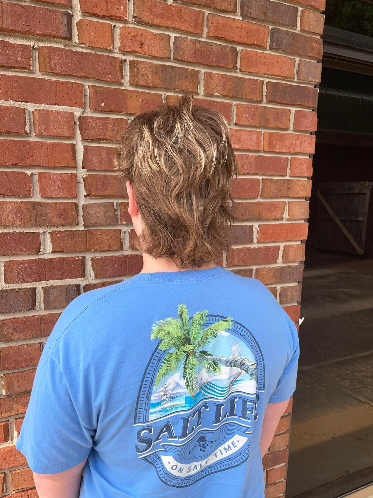 A man wearing a blue shirt with a palm tree on the back is standing in front of a brick wall.