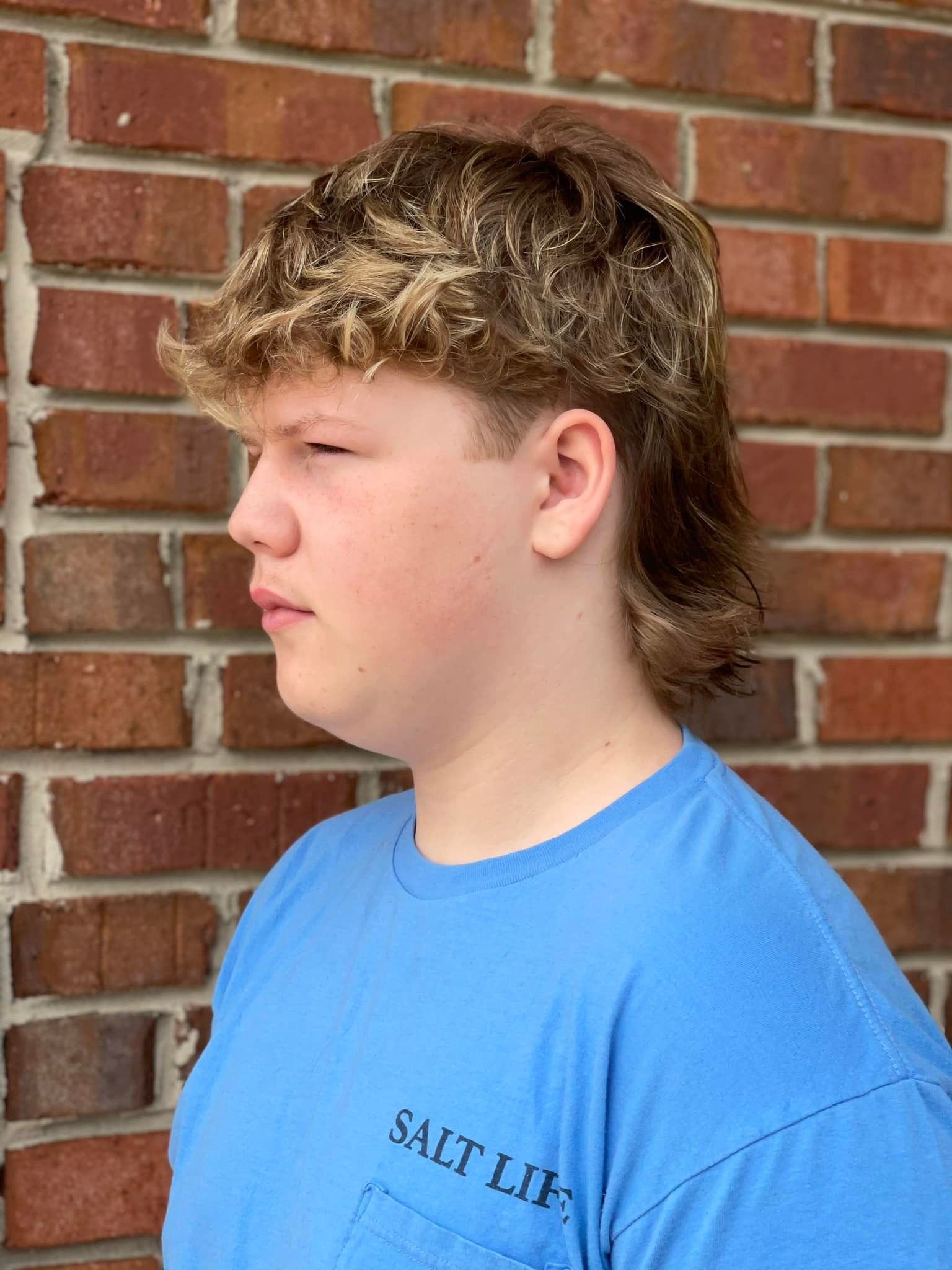 A young man wearing a blue salt life t-shirt is standing in front of a brick wall.