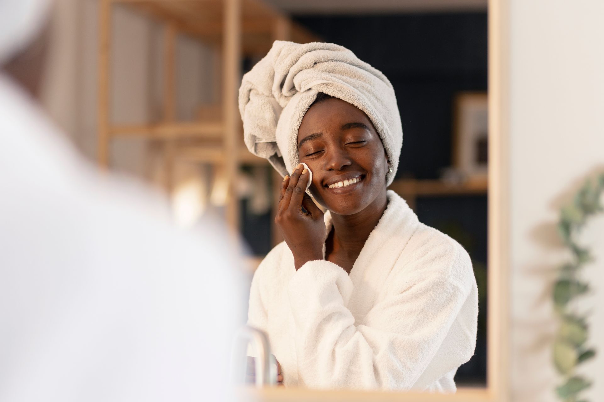 Woman in robe cleanses face with cotton pad, smiling at mirror.