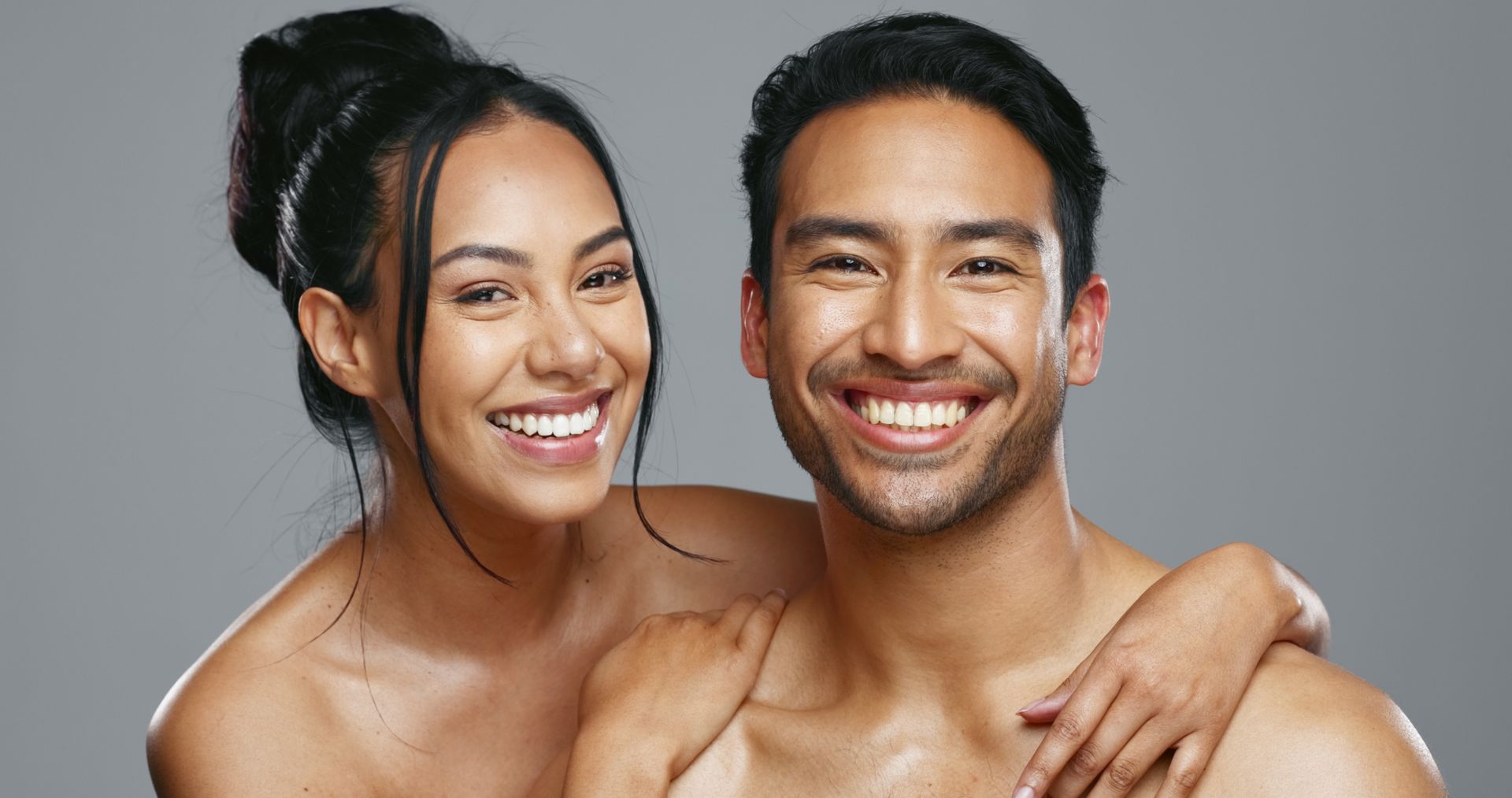 Smiling couple embracing, standing against a gray backdrop. The woman has dark hair up, the man’s hand is on her shoulder.