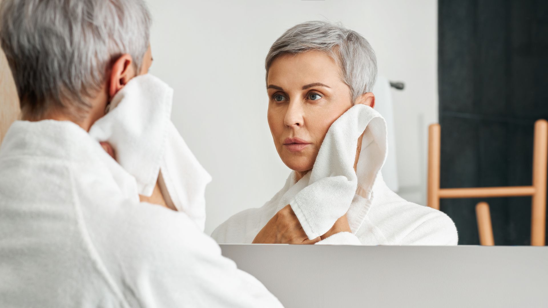 Woman with gray hair in a white robe drying her face with a white towel in front of a mirror.