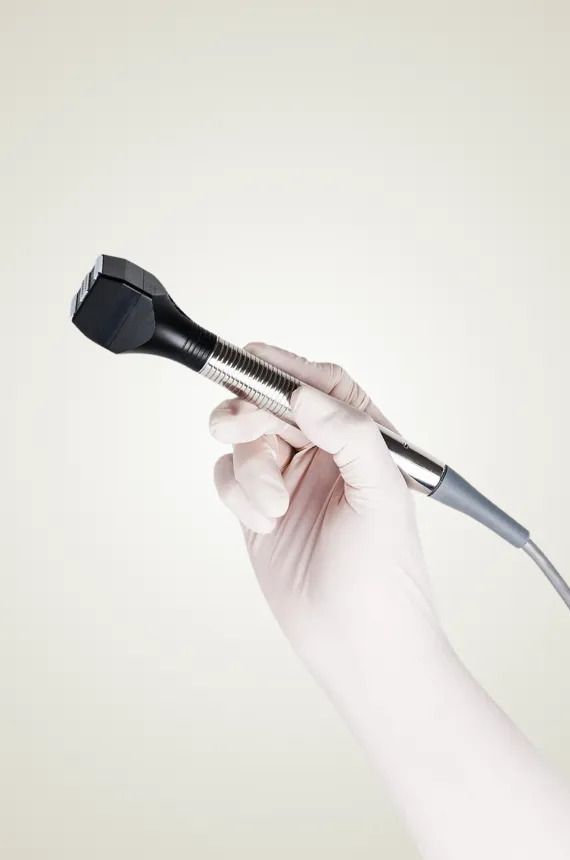 Hand in a white glove holds a medical device, against a pale background.