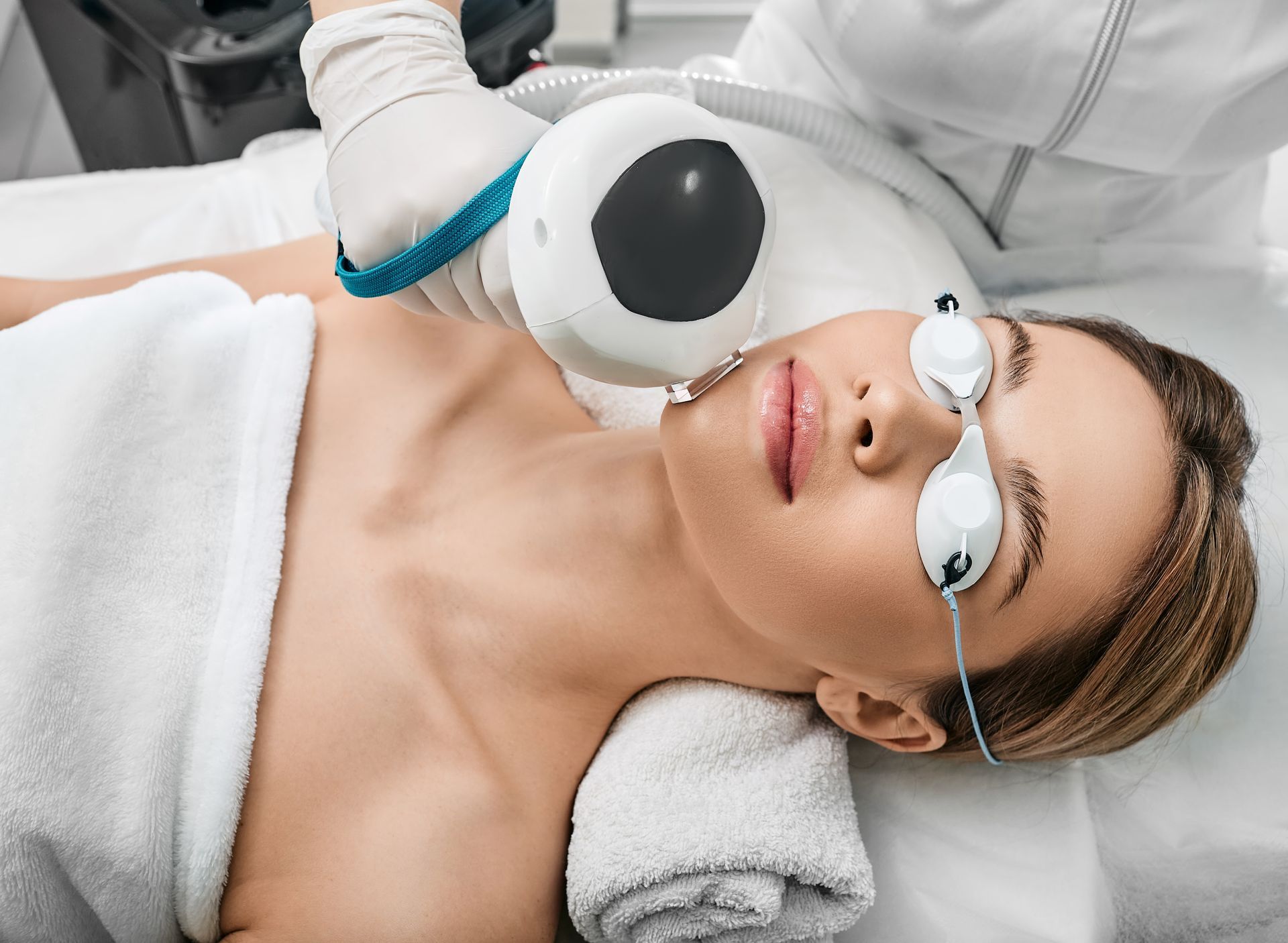 Woman receiving facial laser treatment, lying on a white bed, wearing protective eyewear, white towel.