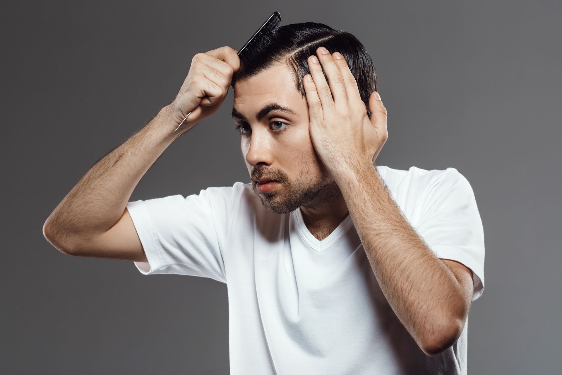 Man combing his dark hair, wearing a white shirt, looking at his reflection. Gray background.