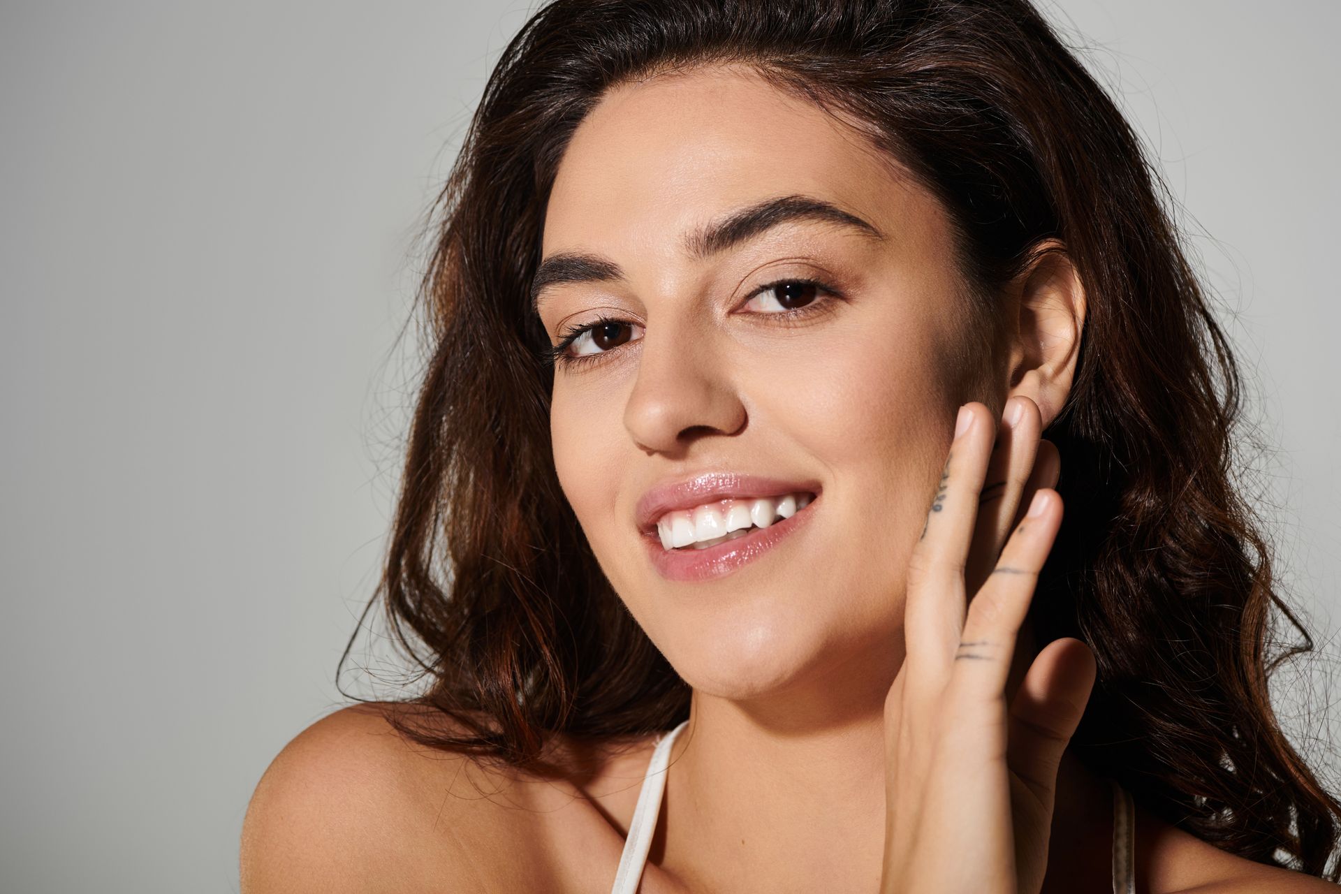Woman with wavy brown hair smiles, touching her cheek. Light skin, white top, against a gray background.
