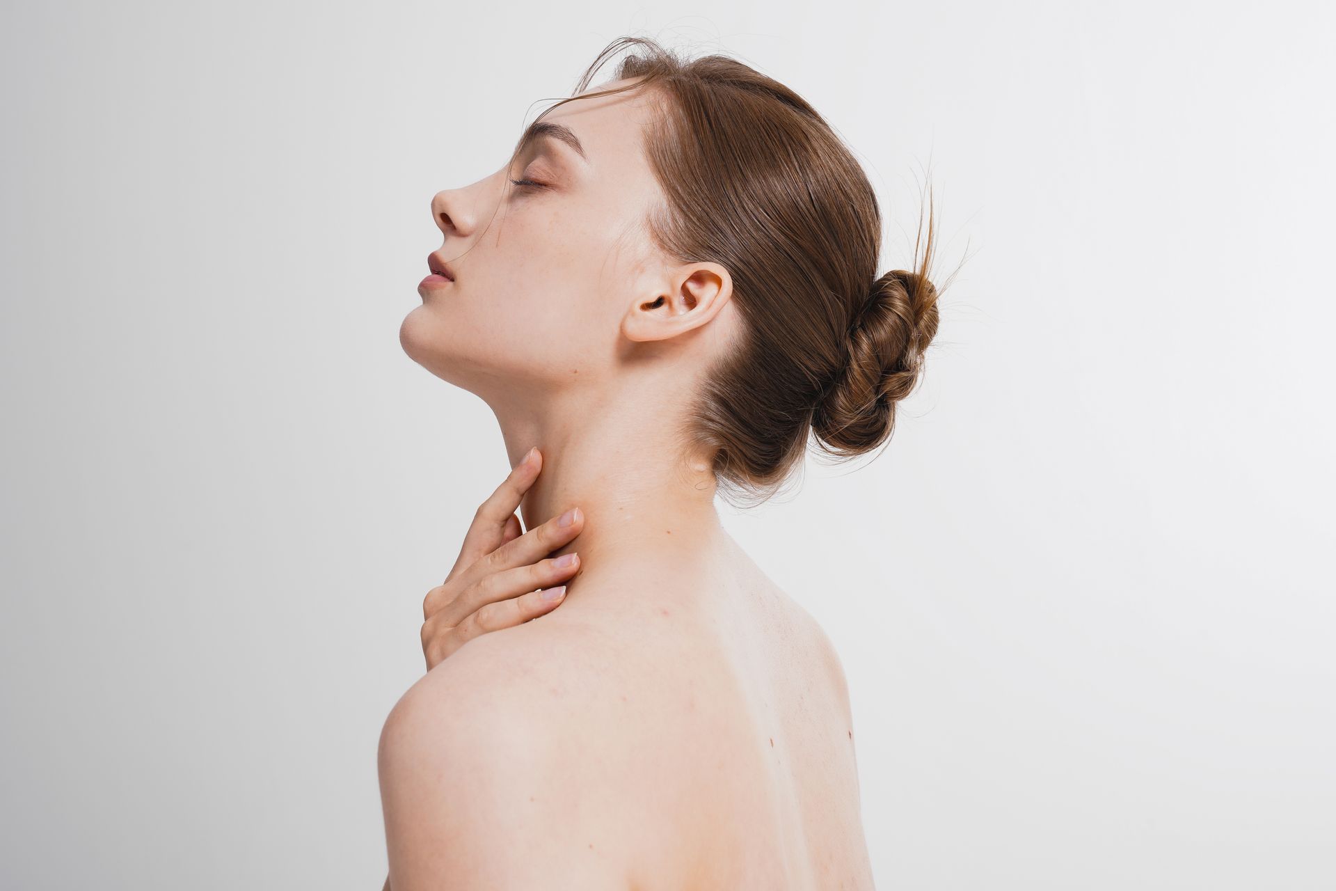 Woman with head tilted back, eyes closed, hand on her neck, bun hairstyle, bare shoulders, white background.
