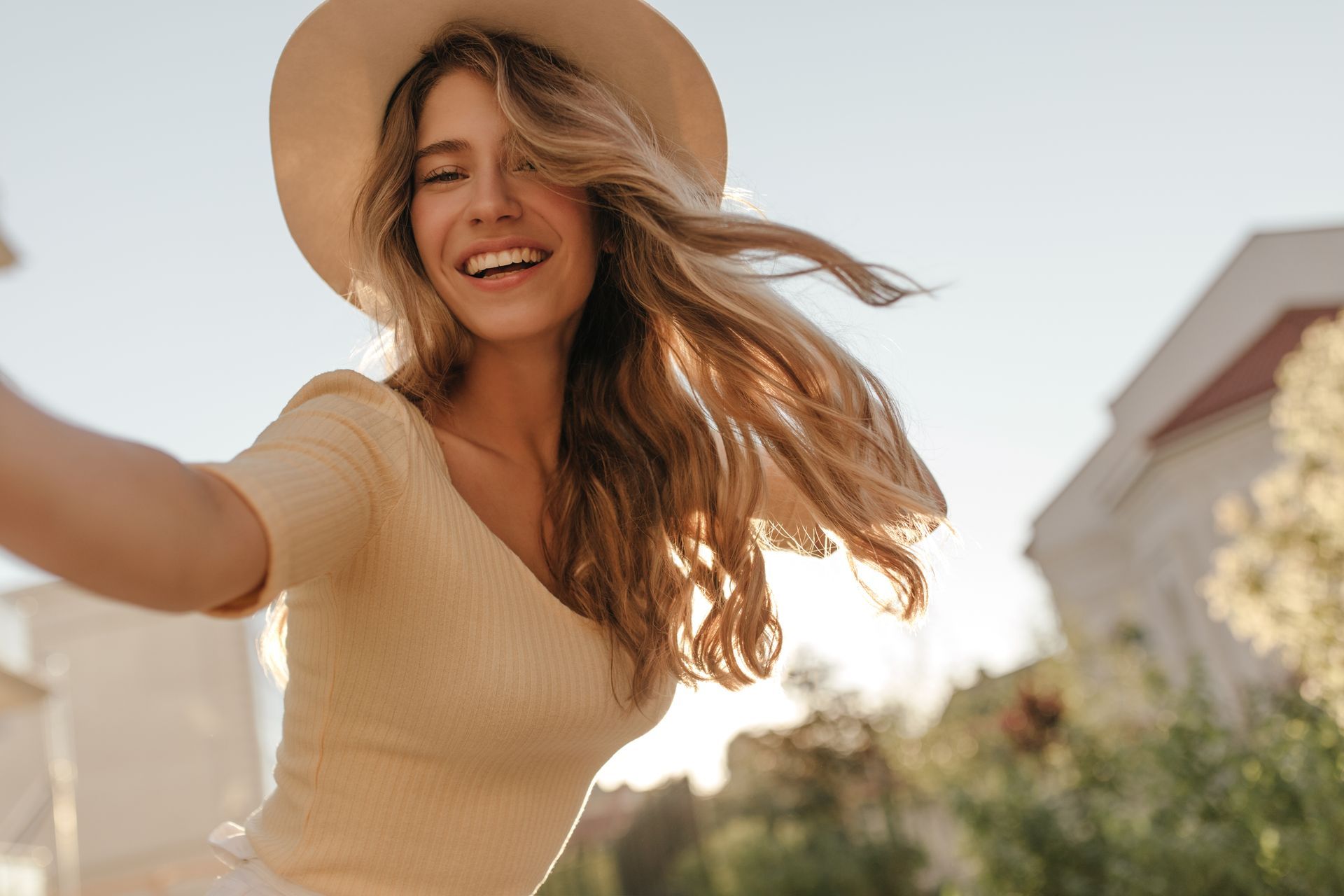 A smiling person wearing a beige top and sun hat outdoors, with wind-blown hair and sunlight in the background.