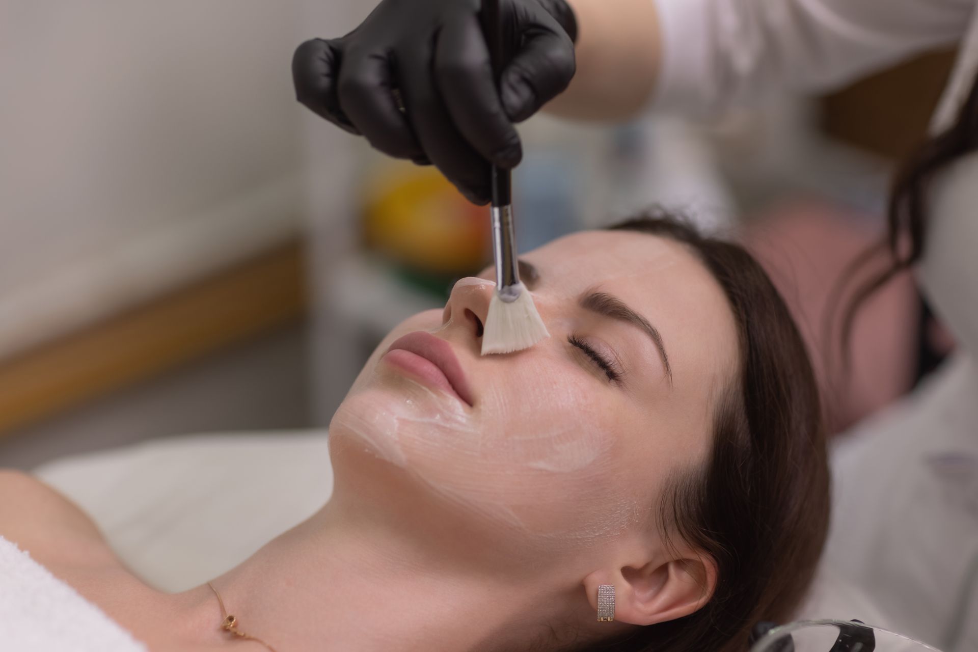 A professional wearing black gloves uses a flat brush to apply a treatment mask to a client's nose in a spa setting.