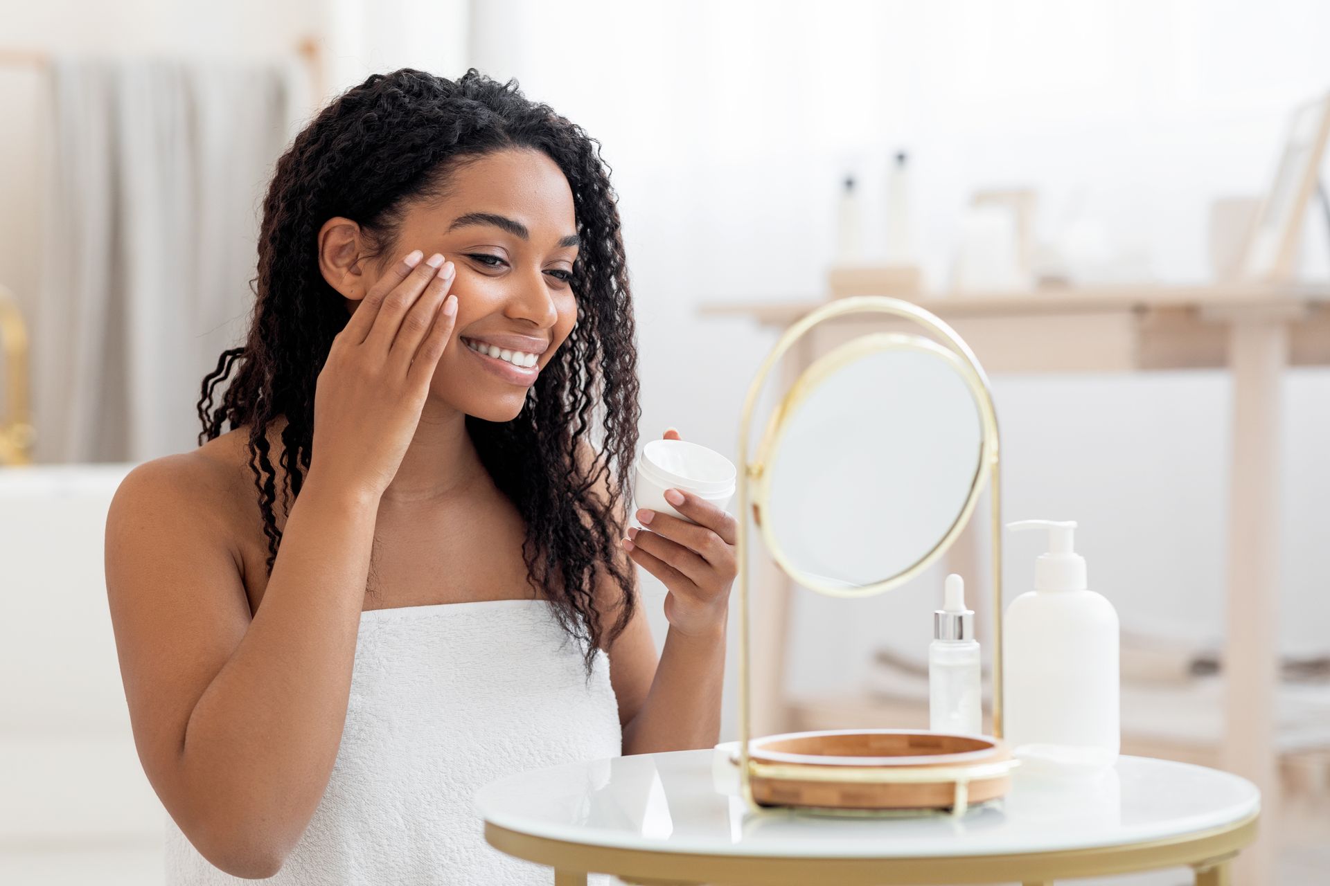 A person wrapped in a white towel applies skincare cream to their cheek while smiling in a bright, modern bathroom.