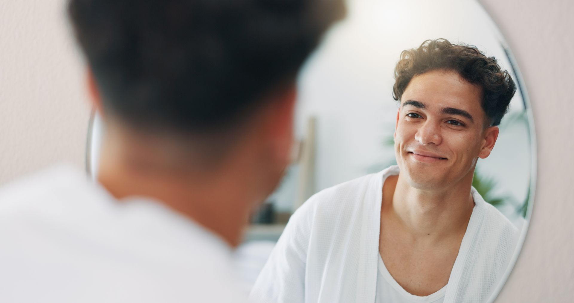 A person with curly hair, wearing a white robe, smiles at their reflection in a circular bathroom mirror.