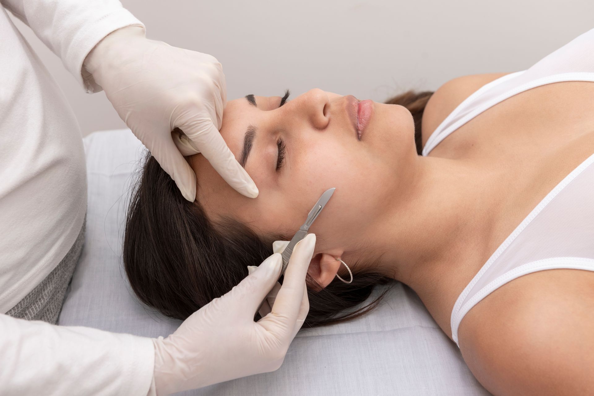 A practitioner wearing white gloves performs a dermaplaning treatment on a client's cheek using a sterile scalpel.
