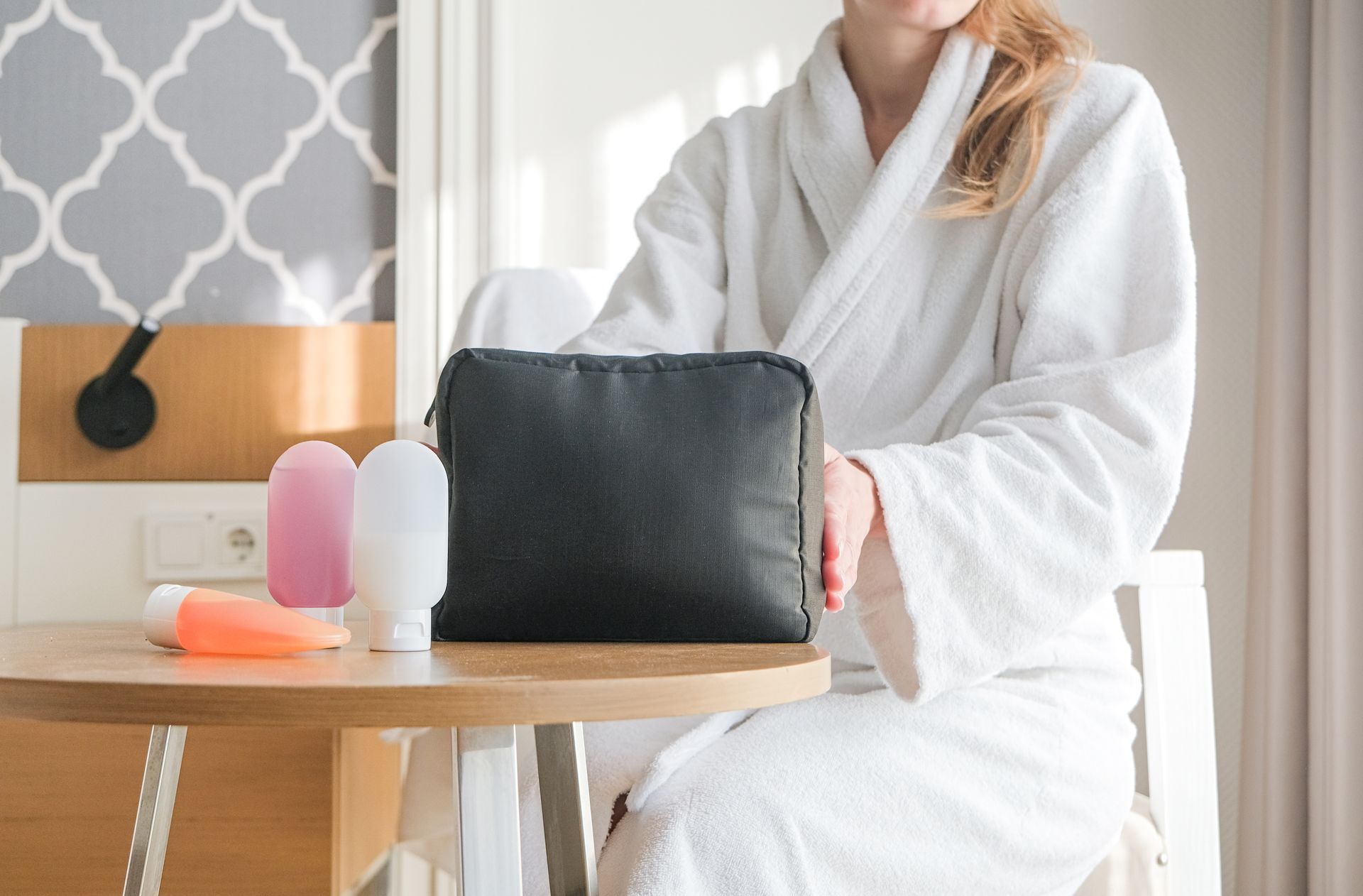 A person in a white bathrobe sits at a wooden table with a black toiletry bag and three travel-sized bottles.