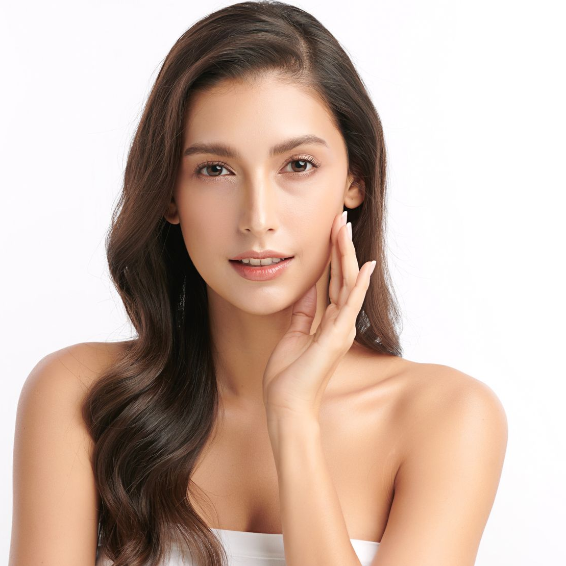 Woman with dark wavy hair and fair skin touches her cheek, smiling, against a white background.