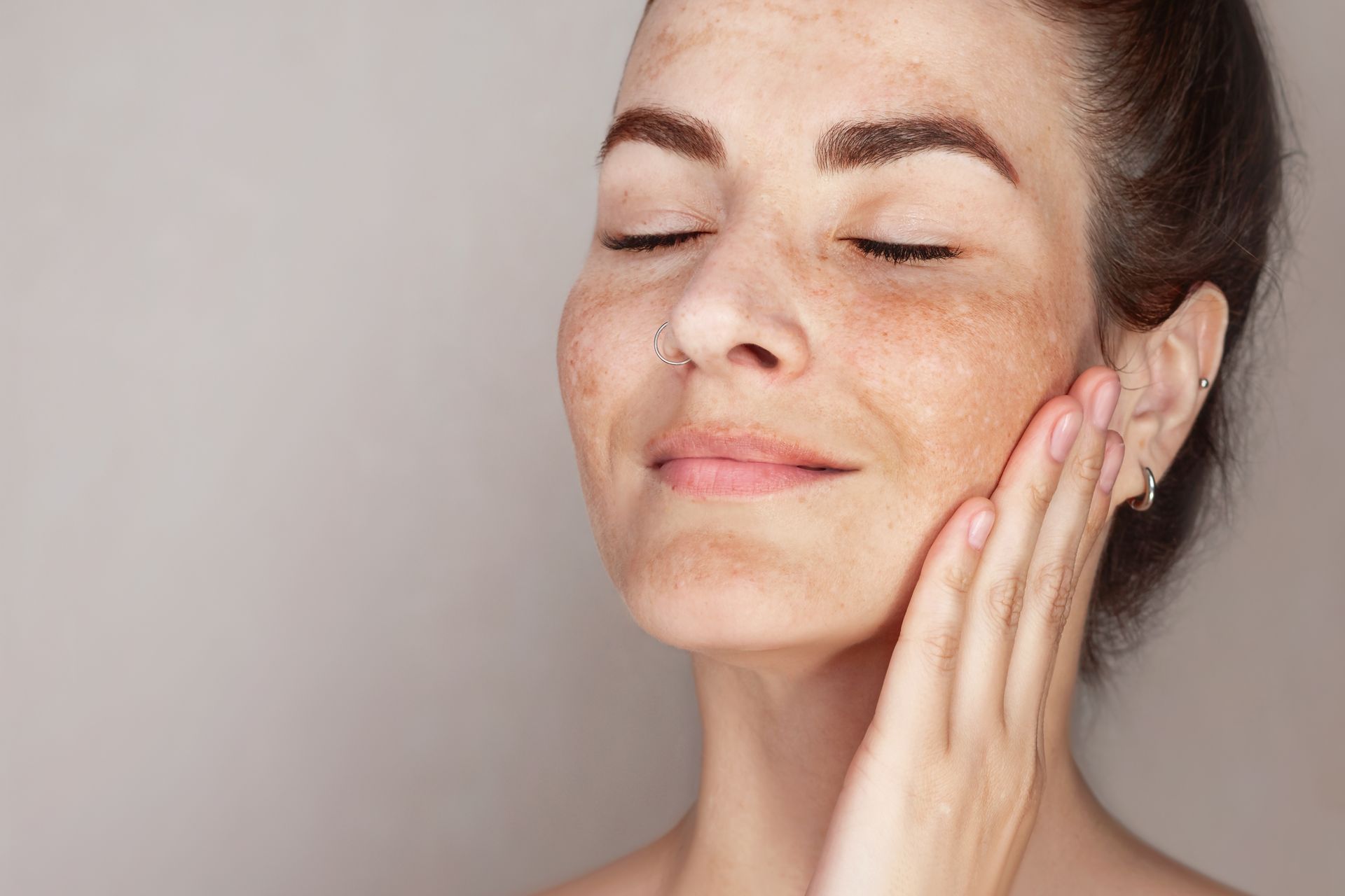 Woman with eyes closed, hand on cheek, displaying hyperpigmentation on face, against a plain backdrop.