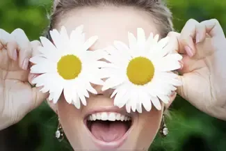 A woman is holding two daisies in front of her eyes.