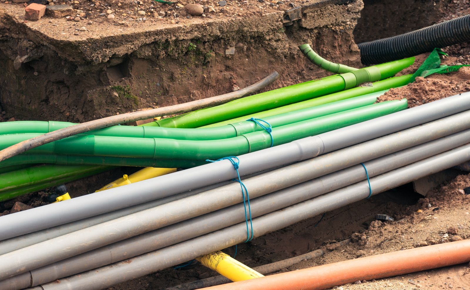 Pipes of varying colors (gray, green, yellow, orange) in a trench, likely for utilities.