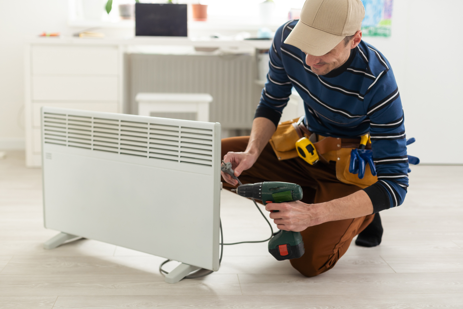Man in work clothes uses a drill to fix a white heater indoors.