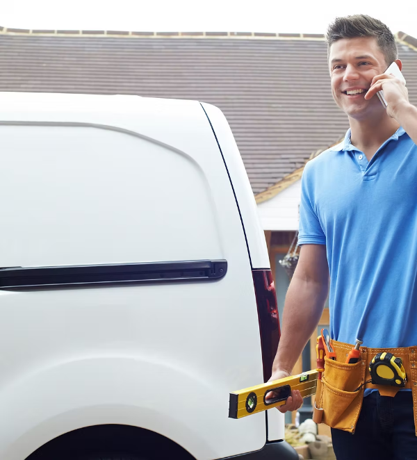 Man in blue shirt, tool belt, talking on phone next to a white van, holding a level.