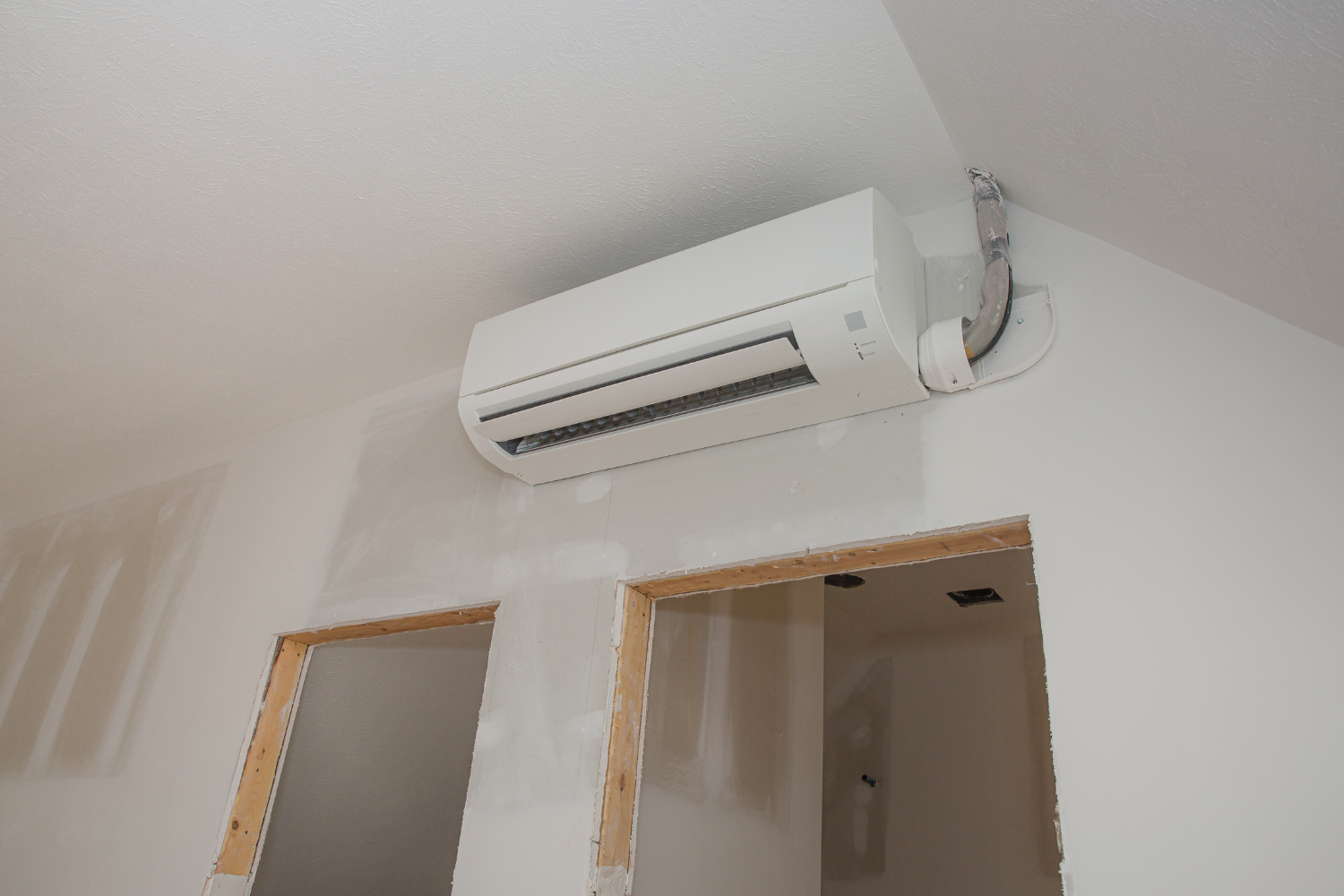 White air conditioning unit mounted on a white wall above two unfinished doorways in a room.