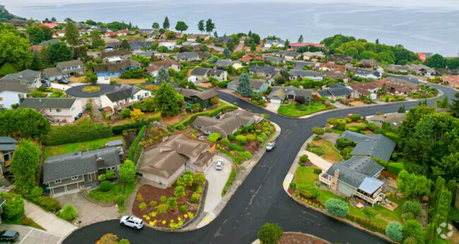 Aerial view of a suburban neighborhood with houses, roads, trees, and a body of water in the background.