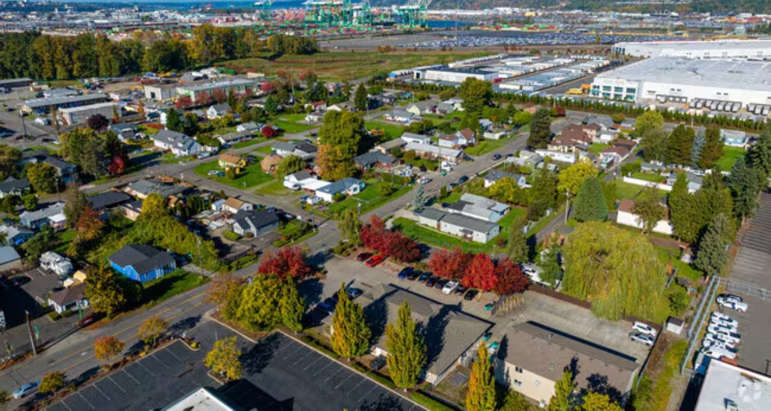 Aerial view of a residential area with trees displaying autumn colors, with a port and industrial area in the background.