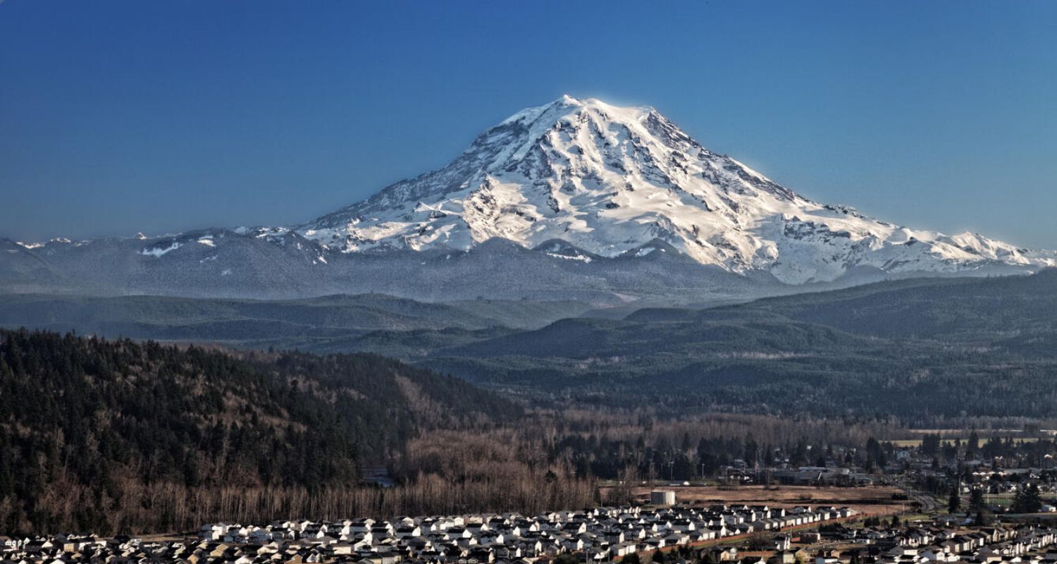 Snow-covered Mount Rainier rises above a valley with houses and trees under a clear blue sky.