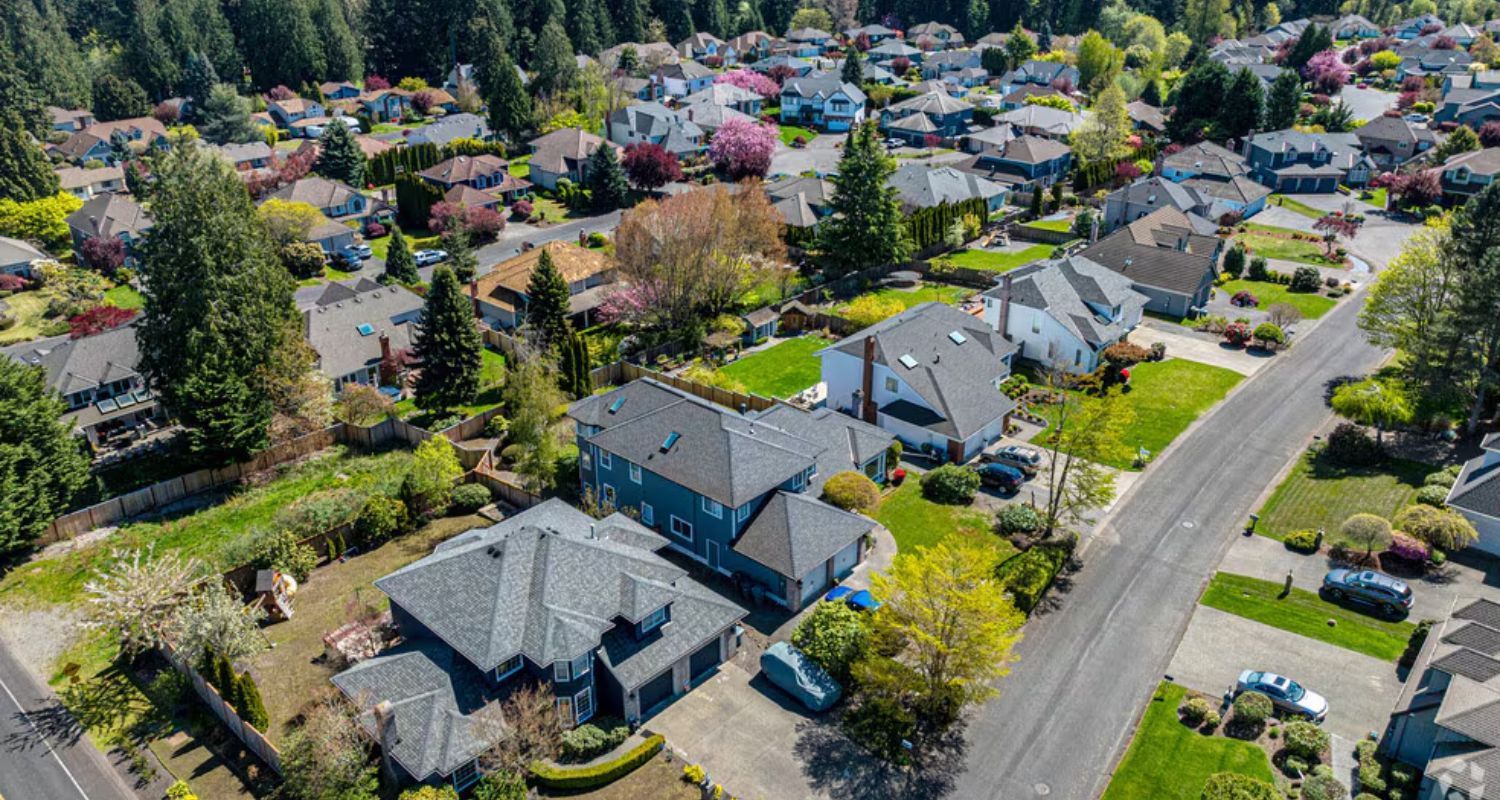 Aerial view of a suburban neighborhood with houses, streets, and trees. Green grass and blue sky.